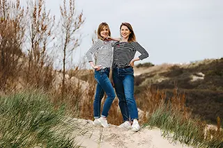 Deux amies posant avec joie sur une dune, vêtues de hauts rayés assortis et de jeans, partageant un moment de complicité dans un paysage de bord de mer naturel.