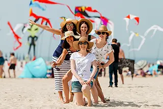 Cinq amies se tiennent bras dessus bras dessous sur la plage, avec des cerfs-volants colorés dans le ciel bleu, symbolisant une journée de célébration et de joie sous le soleil.