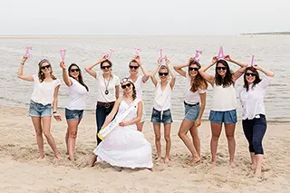 Un groupe d'amies se tient sur la plage avec la future mariée au centre, levant joyeusement des verres en signe de célébration, avec la mer étendue derrière elles.