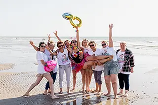 Un groupe hétéroclite célèbre joyeusement sur la plage, portant la future mariée sur les épaules dans une ambiance festive et colorée, avec la mer comme toile de fond.