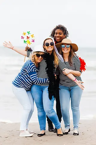 Quatre amies en tenues assorties de marin, partageant un moment complice sur la plage, avec des rires et des étreintes sous un ciel gris, démontrant leur amitié forte.
