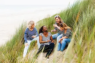 Séance détente pour un groupe d'amies assises dans les herbes des dunes, partageant des histoires et des éclats de rire, avec la plage en arrière-plan.