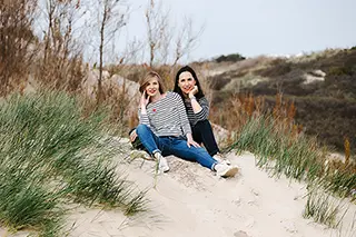 Deux femmes souriantes assises côte à côte sur une dune sablonneuse, entourées d'herbes de dune, reflétant une amitié décontractée et heureuse dans un cadre naturel.