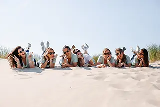 Un groupe de femmes allongées sur le sable forme une ligne oblique, leurs expressions détendues et les lunettes de soleil évoquant un parfait après-midi de farniente.