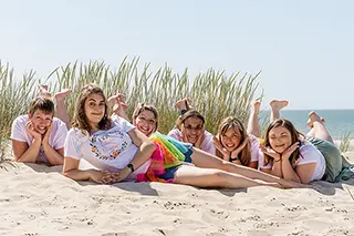 Cinq femmes souriantes se prélassent sur les dunes, profitant d'un moment de détente lors d'une journée ensoleillée, l'amitié et le sable fin à l'unisson.