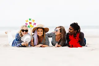 Allongées sur le sable, quatre amies profitent d'un moment de détente à la plage, partageant rires et conversations légères, avec des accessoires festifs pour agrémenter leur journée.
