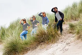 Un groupe d'amies en marinières explore les dunes, leurs poses indiquant une promenade dynamique et enjouée, avec un fond de verdure et de sable fin.