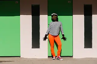 Une femme à la mode posant avec confiance devant une cabine de plage verte et blanche, vêtue d'un pantalon orange vif, d'un haut rayé et coiffée d'un chapeau tendance, complétant son style avec des lunettes de soleil et des baskets.