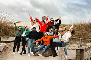 Un groupe d'amies levant joyeusement les bras en l'air, partageant un moment de bonheur collectif devant les hautes herbes des dunes de sable.