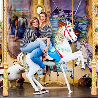 Sur un carrousel aux couleurs vives, une amie rayonne de bonheur, perchée sur une girafe de manège, saisissant un moment de liberté et d'enchantement.
