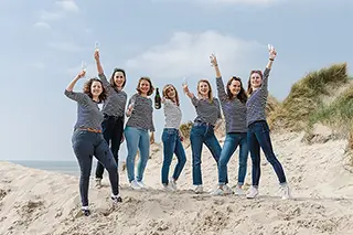 Un groupe de sept femmes joyeuses sautant ensemble sur une plage de sable, les bras levés en l'air, avec des dunes en arrière-plan sous un ciel légèrement nuageux.