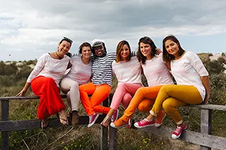 Assises sur une barrière en bois au milieu des dunes, six femmes rayonnent de bonheur en t-shirts blancs et pantalons colorés, partageant des sourires et de la bonne humeur.