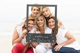 Un groupe de femmes souriantes encadre leur amie dans un cadre symbolique portant l'inscription 'Team Bride', capturant la joie et l'anticipation d'un mariage imminent.