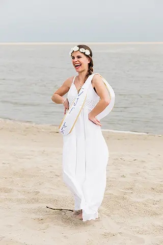 Une amie en robe blanche et couronne de fleurs incarne la joie et l'élégance sur la plage, son sourire éclatant reflétant la célébration et l'anticipation d'un moment spécial.