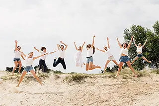 Dans un élan de bonheur, un groupe d'amies saute en l'air sur les dunes, capturant un moment de légèreté et d'excitation avec le ciel nuageux en arrière-plan.